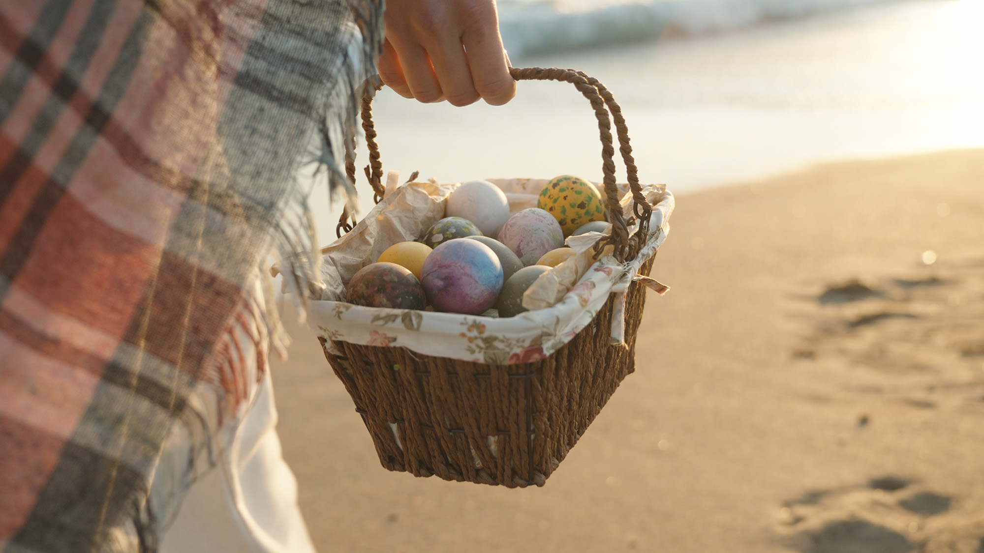 A young woman walks along the beach at sunset carrying a basket of Easter eggs. I’m walking towards the sun, the sea waves are behind me.